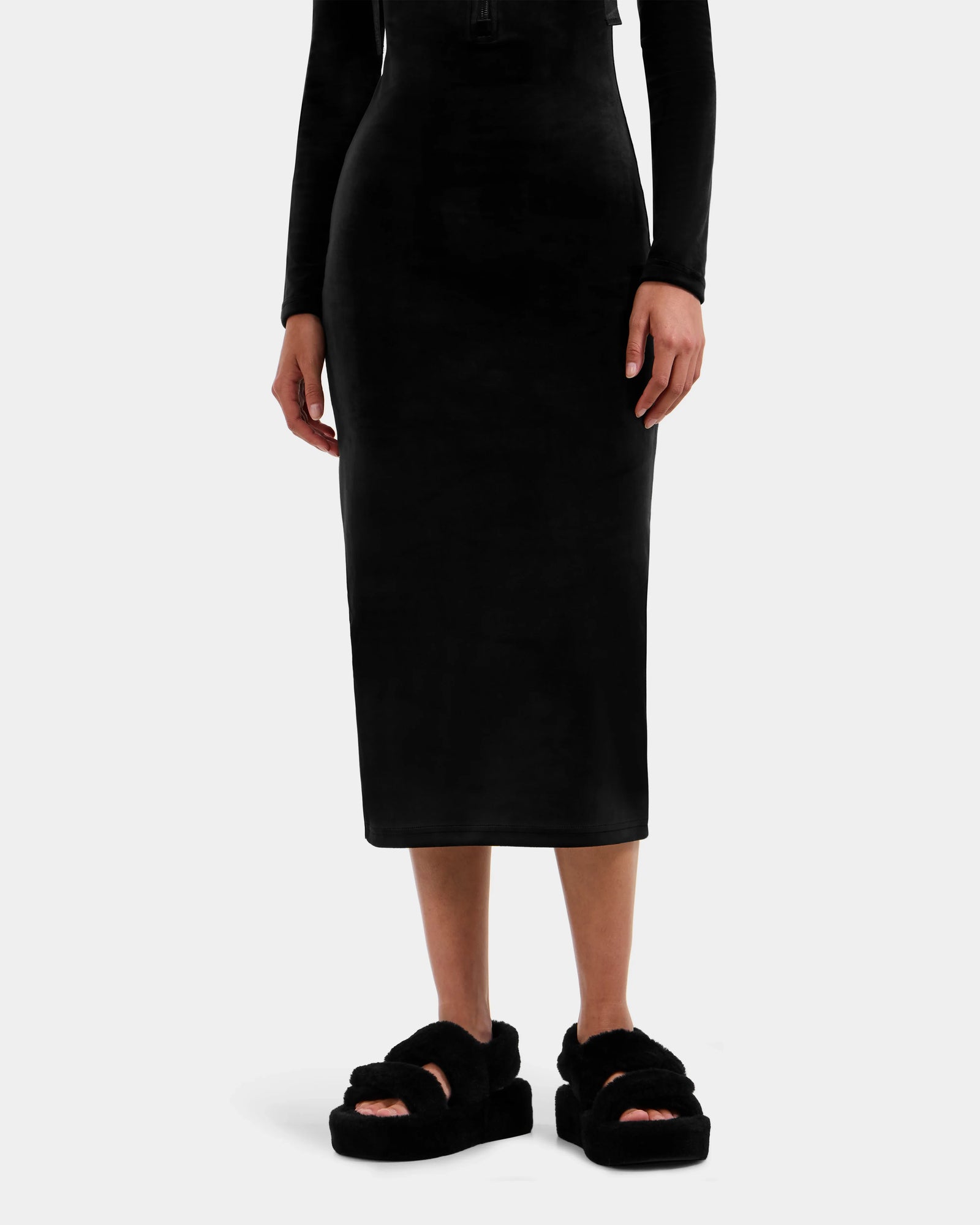 A person showcases the lower half in Rudsak's ISABELLA - L525502 Black fitted, long-sleeve midi dress with black fuzzy platform sandals, standing against a plain white background and highlighting the brand's sleek style.