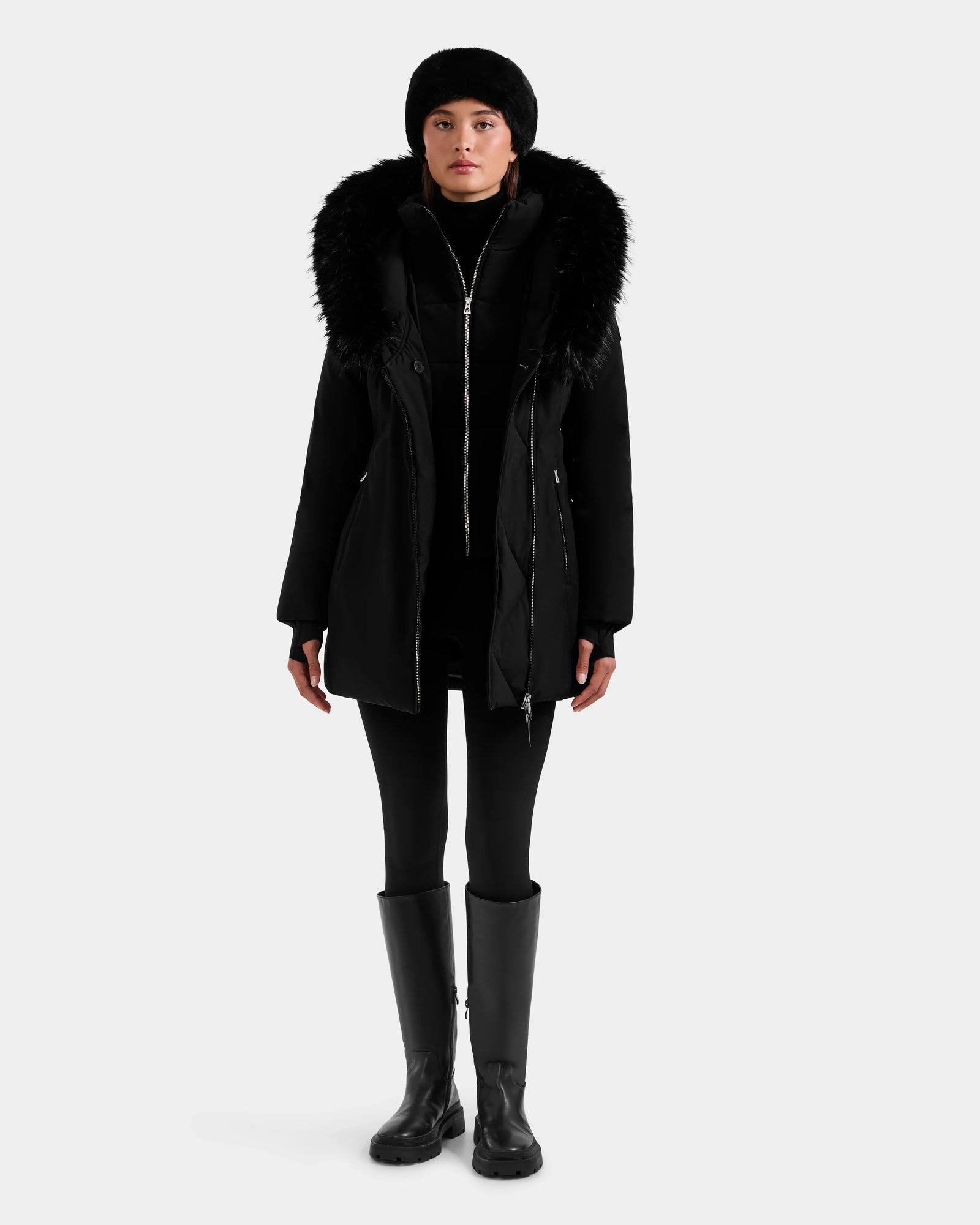 A person stands facing forward against a white background, wearing the Rudsak ISELLE - L125547 Black women’s down parka with a fur hood, styled with a black hat, black leggings, and black knee-high boots.