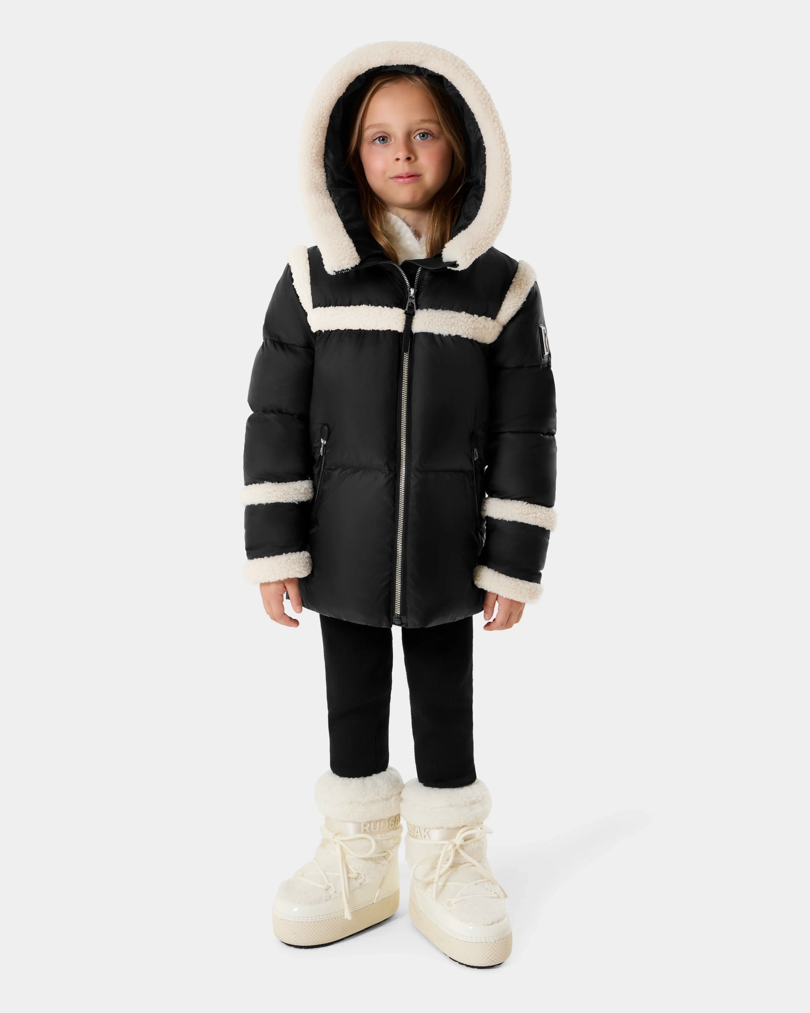 A young child wears the Rudsak JULIANA K - K125550 Black puffer jacket with white fur trim and hood, paired with black pants and white fur-lined boots, standing against a plain light gray background.