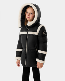 A young girl wears the Rudsak JULIANA K - K125550 Black winter coat with white faux fur trim, standing against a light gray background and gazing slightly upward and to the side.