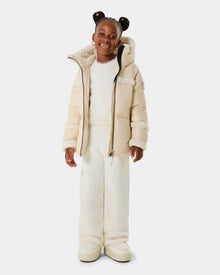 A young girl smiles wearing the Rudsak JULIANA K - K125550 Vanilla puffer jacket over a white fuzzy outfit with matching wide-leg pants and platform shoes, her hair in two buns, standing against a plain white background.