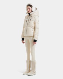A woman models the Rudsak JULIANA - L124509 Vanilla short down puffer with matching earmuffs, fitted pants, and tall winter boots against a white backdrop—a stylish must-have for any winter wardrobe.