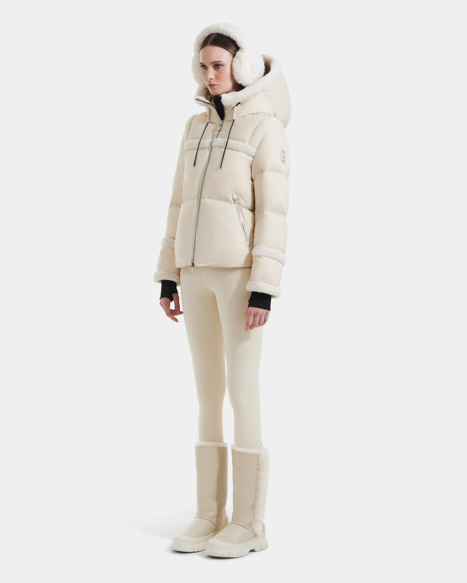 A woman models the Rudsak JULIANA - L124509 Vanilla short down puffer with matching earmuffs, fitted pants, and tall winter boots against a white backdrop—a stylish must-have for any winter wardrobe.