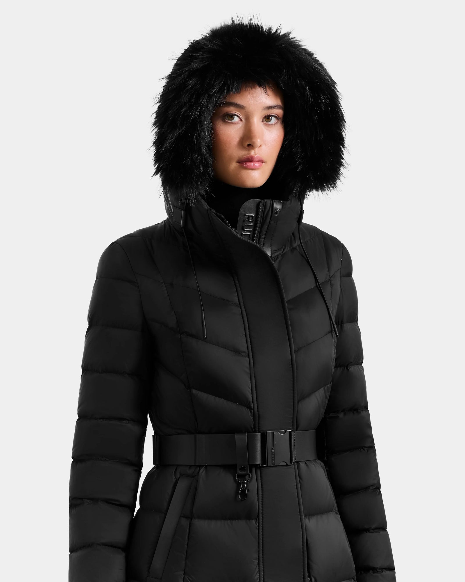 A person wearing the Rudsak JULIETA M-L - L125559 Black belted maxi down puffer coat with a large faux fur hood and weather-resistant shell poses against a light gray background.