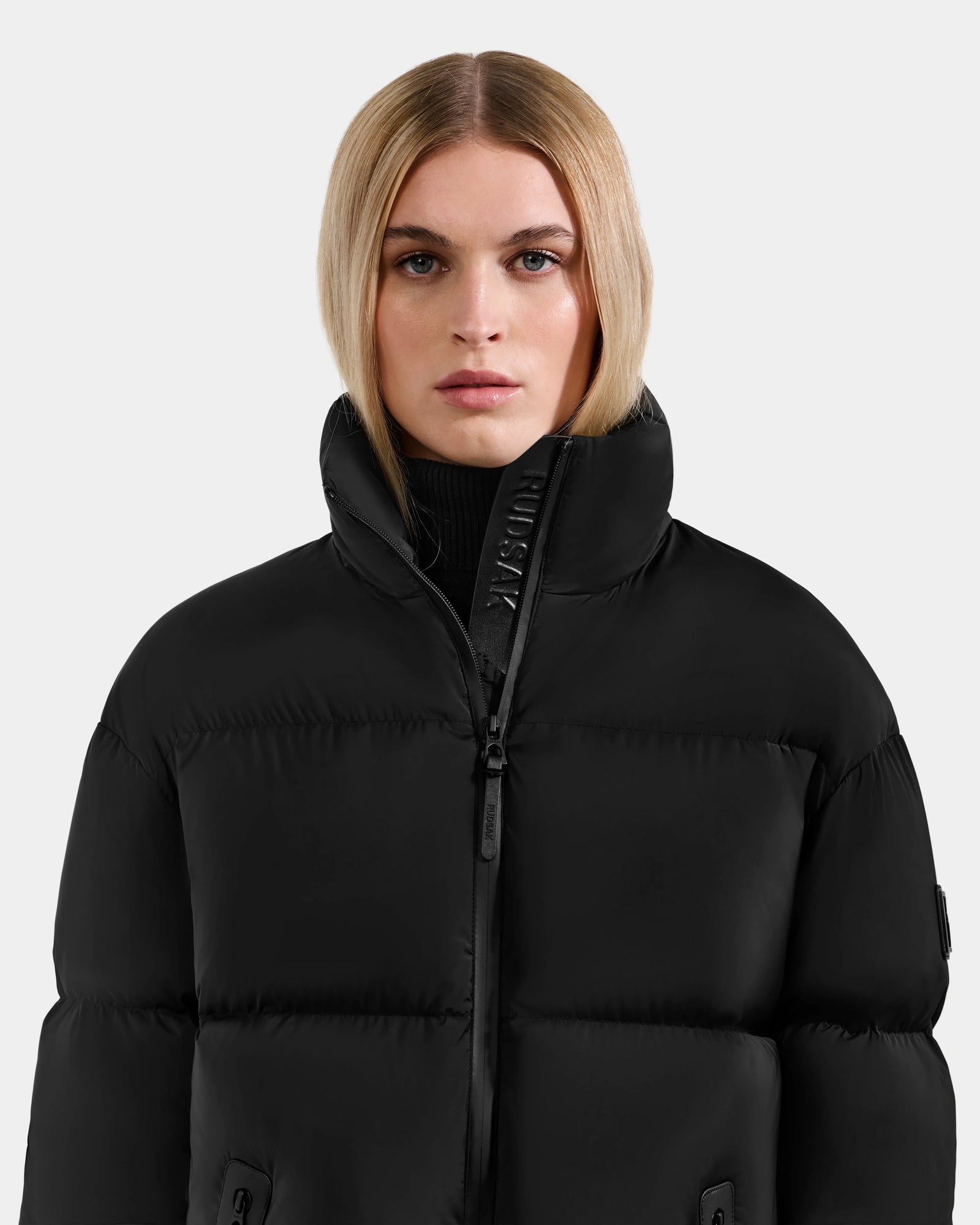 A woman with straight blonde hair wears the Rudsak KARLEE M - 8125681 Black women’s down puffer jacket, zipped to the collar, and stands against a plain light gray background, looking directly at the camera with a neutral expression.