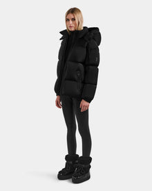 A woman with straight blonde hair stands slightly turned, wearing the Rudsak KARLEE M - 8125681 Black down puffer jacket, black leggings, and black winter boots against a plain white background.