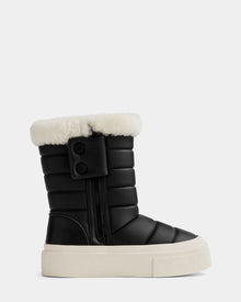 The Rudsak KATHIE - L225516 Black is a women’s black quilted mid boot in vegan leather with a thick white sole, side zipper, two black snap buttons, and plush white faux fur lining at the top. Light gray background.