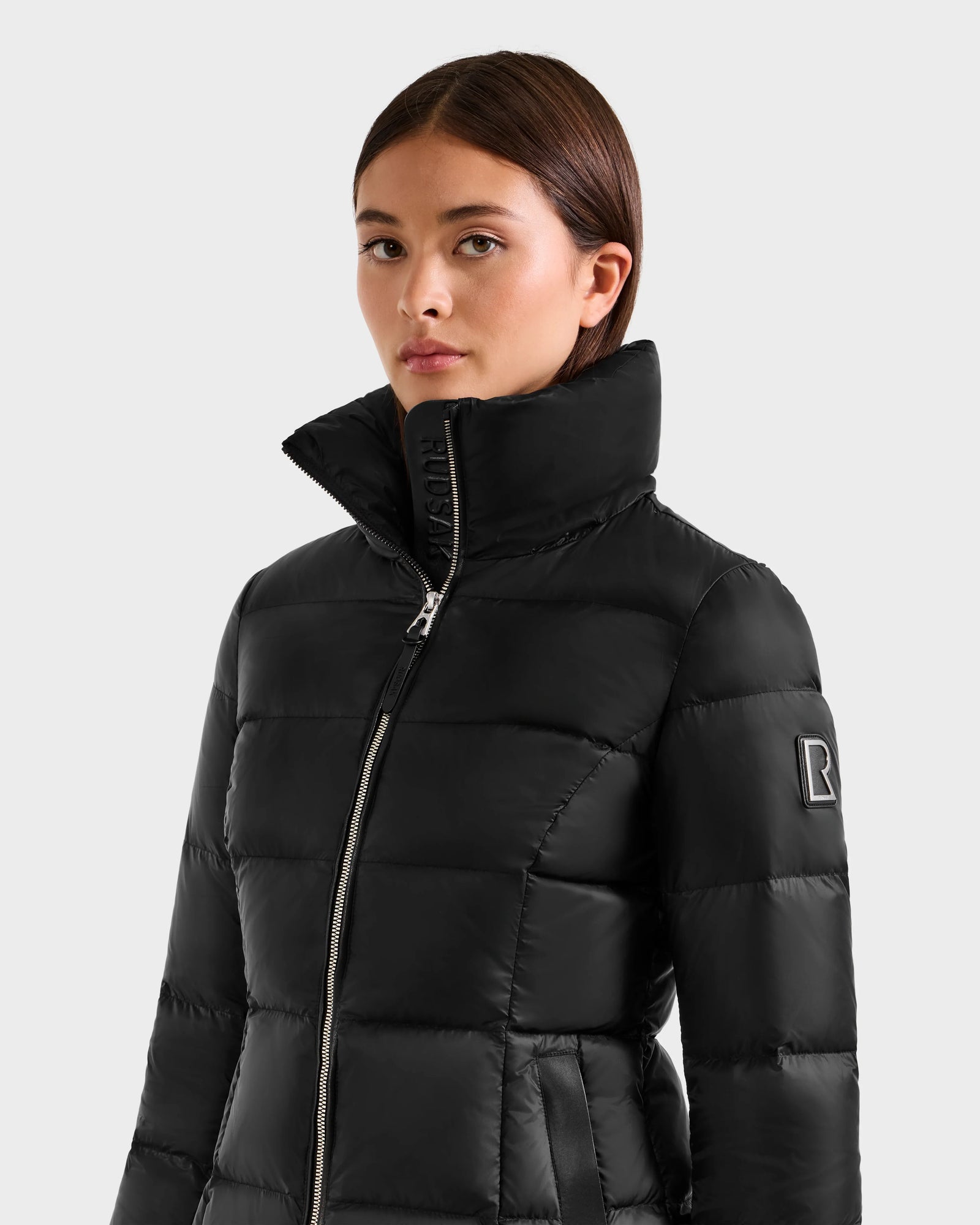 A woman wears the Rudsak KATIA - L125564 Black weather-resistant puffer jacket with a high collar, front zipper, and an "R" patch on the upper sleeve, standing against a light gray background. Ideal as an everyday puffer.