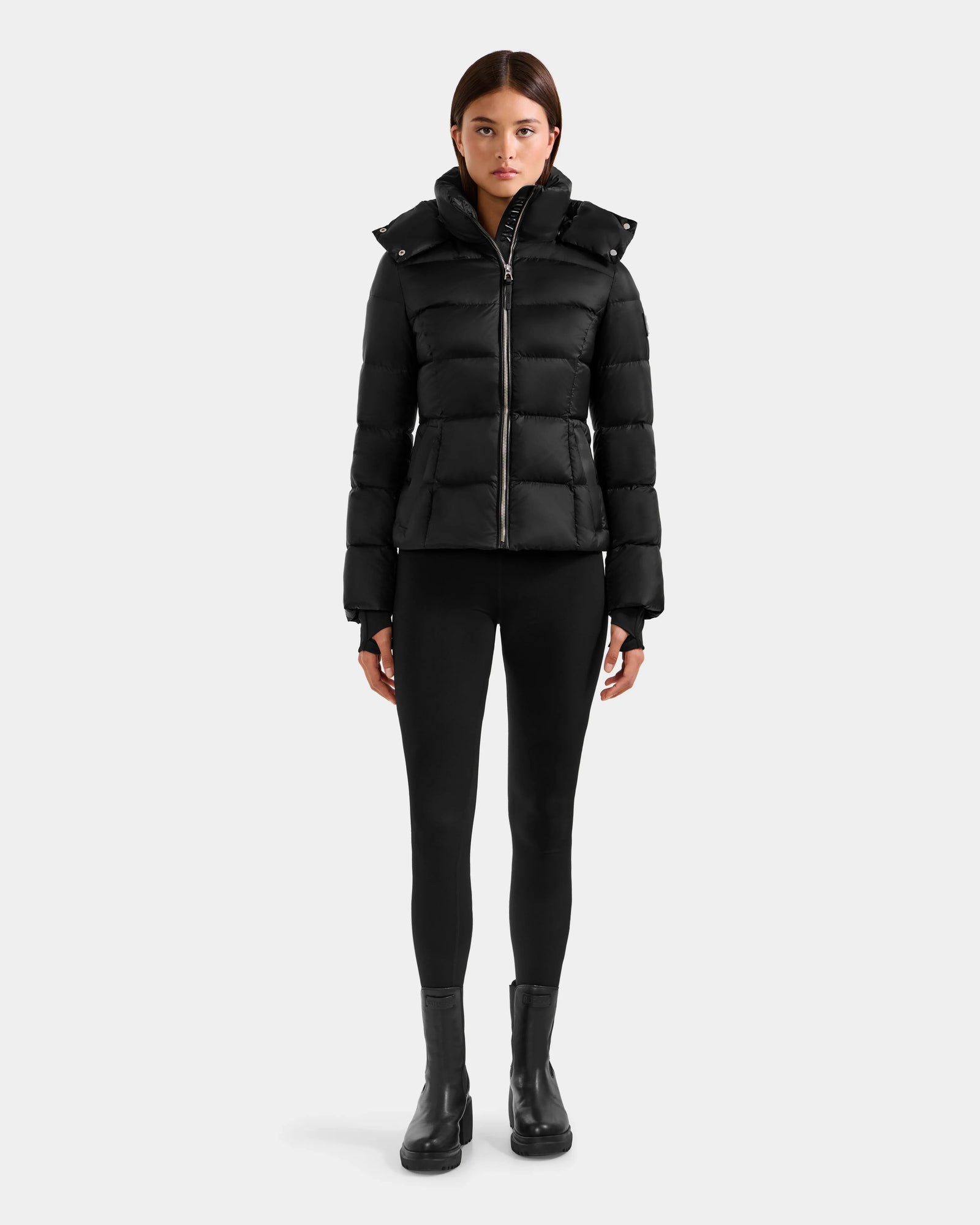 A person stands facing forward in a Rudsak KATIA - L125564 Black weather-resistant puffer jacket, black leggings, and black ankle boots against a plain light gray background.