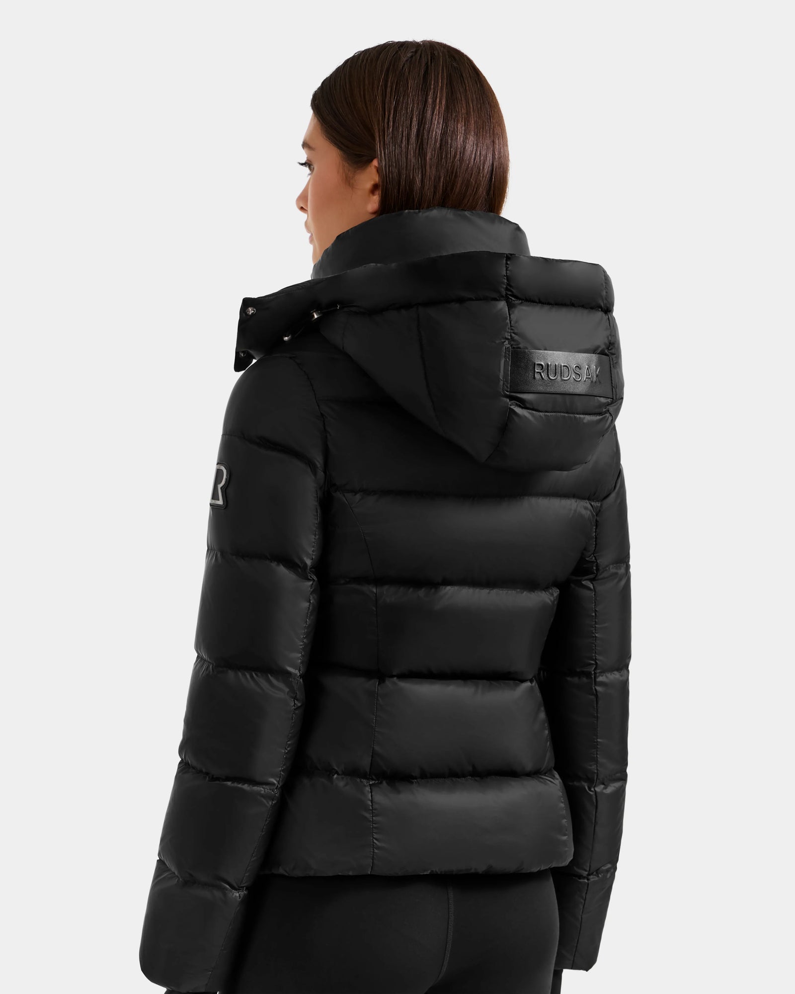 A woman with straight brown hair wears the Rudsak KATIA - L125564 Black, a weather-resistant, quilted puffer jacket with a hood and logo details on the upper back and left sleeve, standing with her back to the camera.