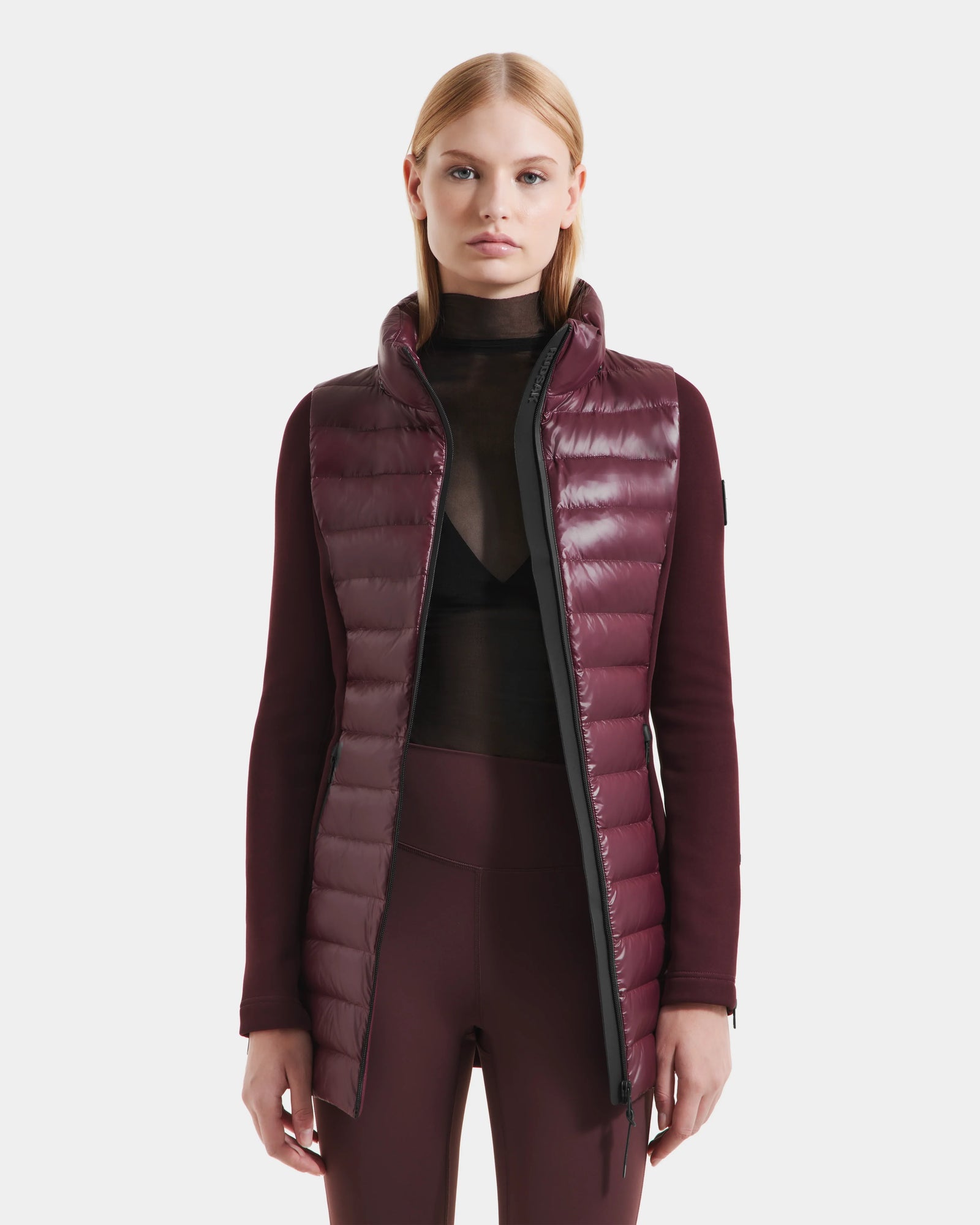 A woman with straight blonde hair wears the Rudsak MARRY S - L125521 Wine weather-resistant quilted vest over a sheer black top and maroon leggings, standing against a plain white background.