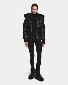 A person stands facing forward wearing the Rudsak MAUDE Q-S - L124553 Black women’s quilted down bomber with a fur-trimmed hood, styled with black leggings, boots, and turtleneck against a plain white background.