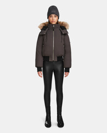 A woman faces forward against a white background, wearing the Rudsak MAUDE - L124522 Grey jacket—a dark brown puffer with premium down and fur-lined hood—paired with a gray beanie, black leggings, and black boots.