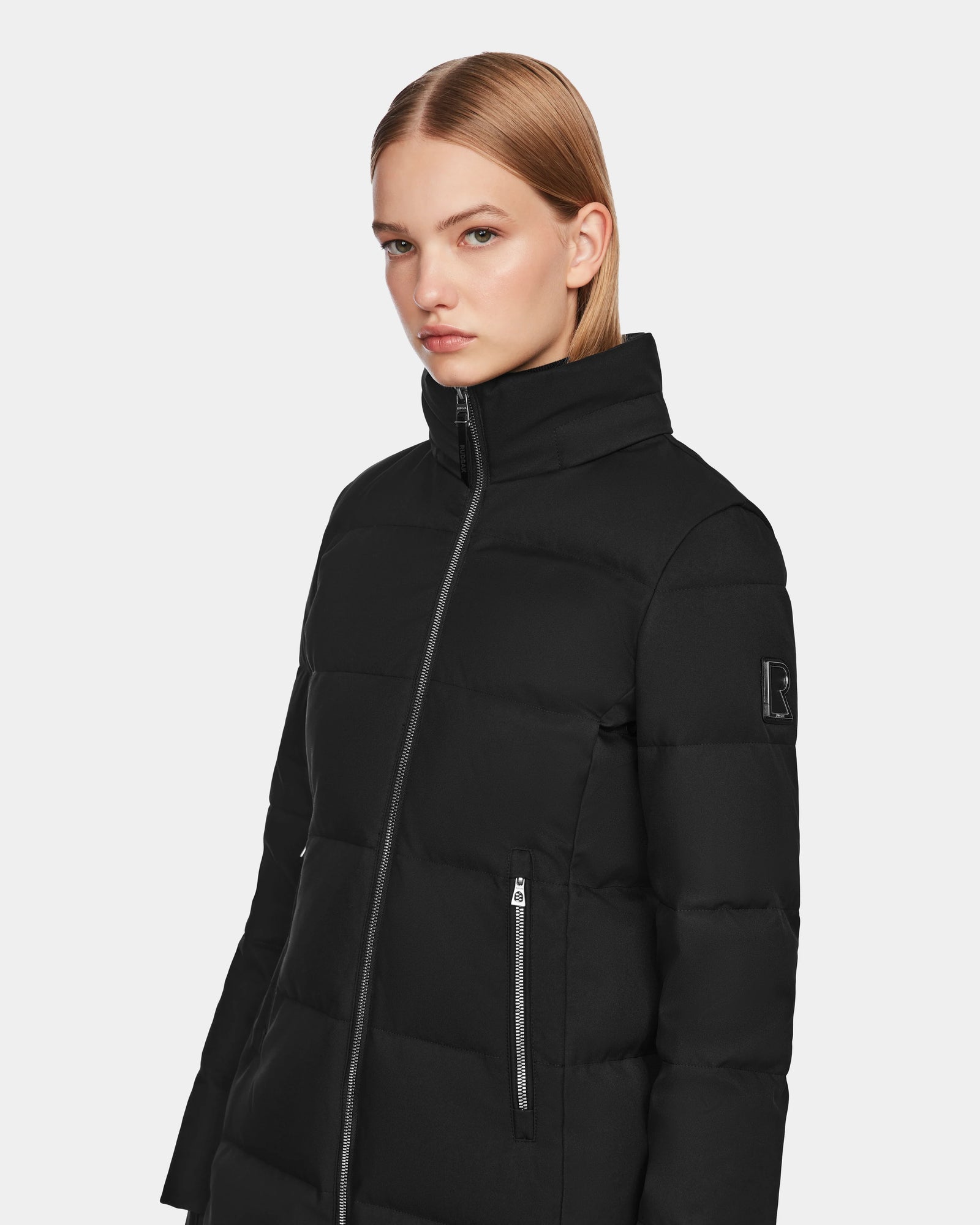 A woman with straight blonde hair models the Rudsak MIRIAM - L123544 Black womens down puffer jacket, featuring a high collar and zippered pockets, against a plain light background.