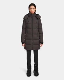 A woman stands facing forward, wearing the Rudsak MIRIAM - L123544 Grey womens down puffer coat with a hood, paired with black leggings and winter boots against a plain white background—a stylish pick for winter.