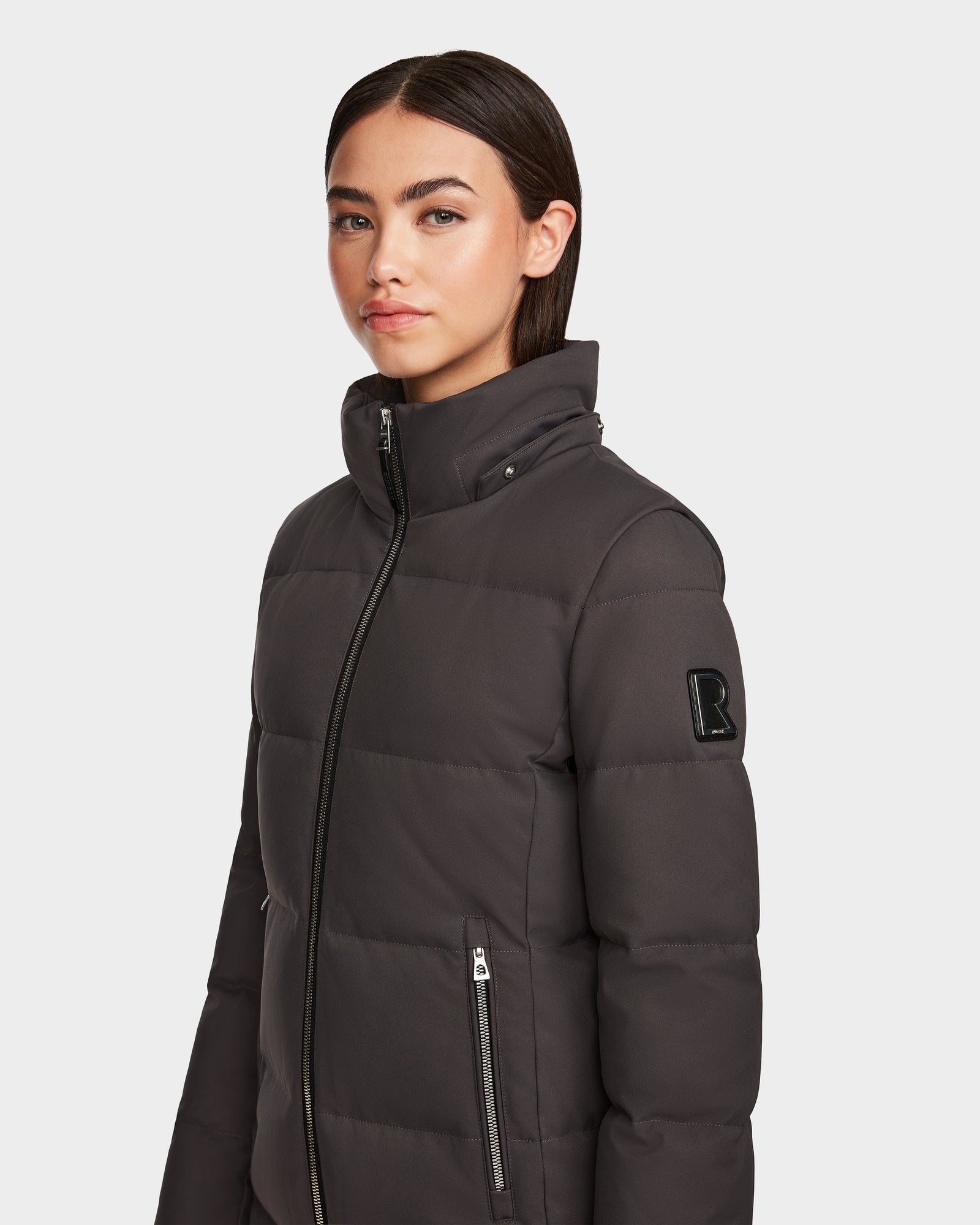 A woman with straight dark hair wears the Rudsak MIRIAM - L123544 Grey womens down puffer jacket, featuring a high collar, zipped pockets, and a black R patch on the sleeve. She stands against a plain white background.