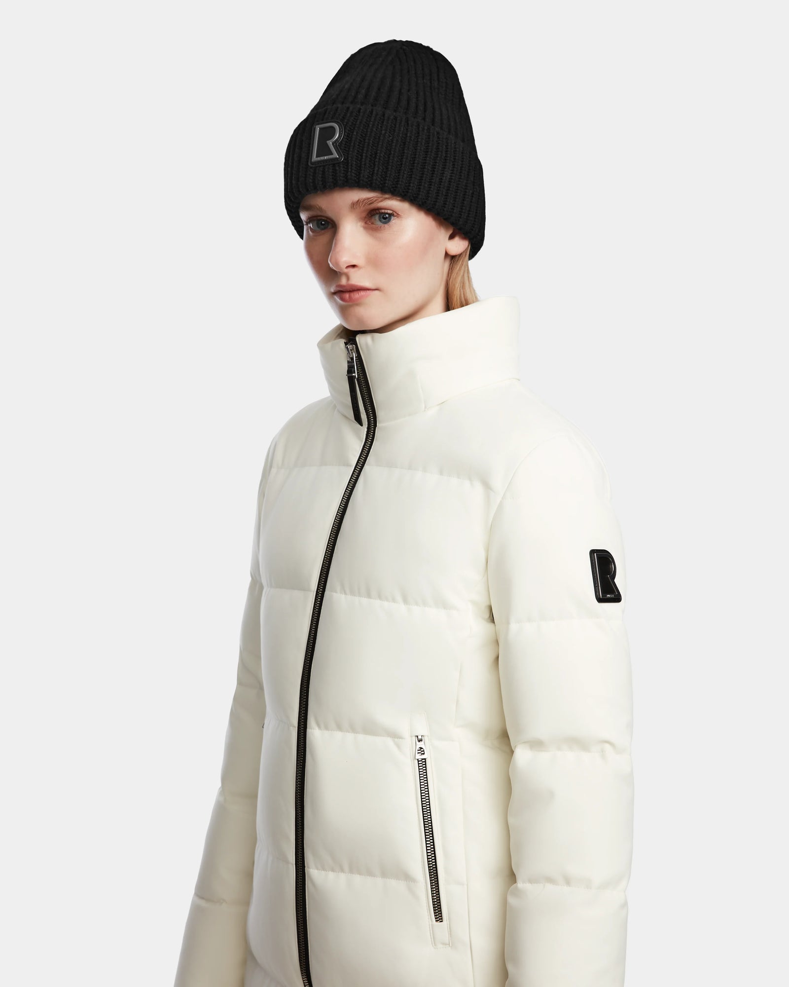 A woman wears the Rudsak MIRIAM - L123544 Iceland women’s puffer jacket with a high collar and zippered pocket, paired with a black logo knit beanie, standing against a plain light background.