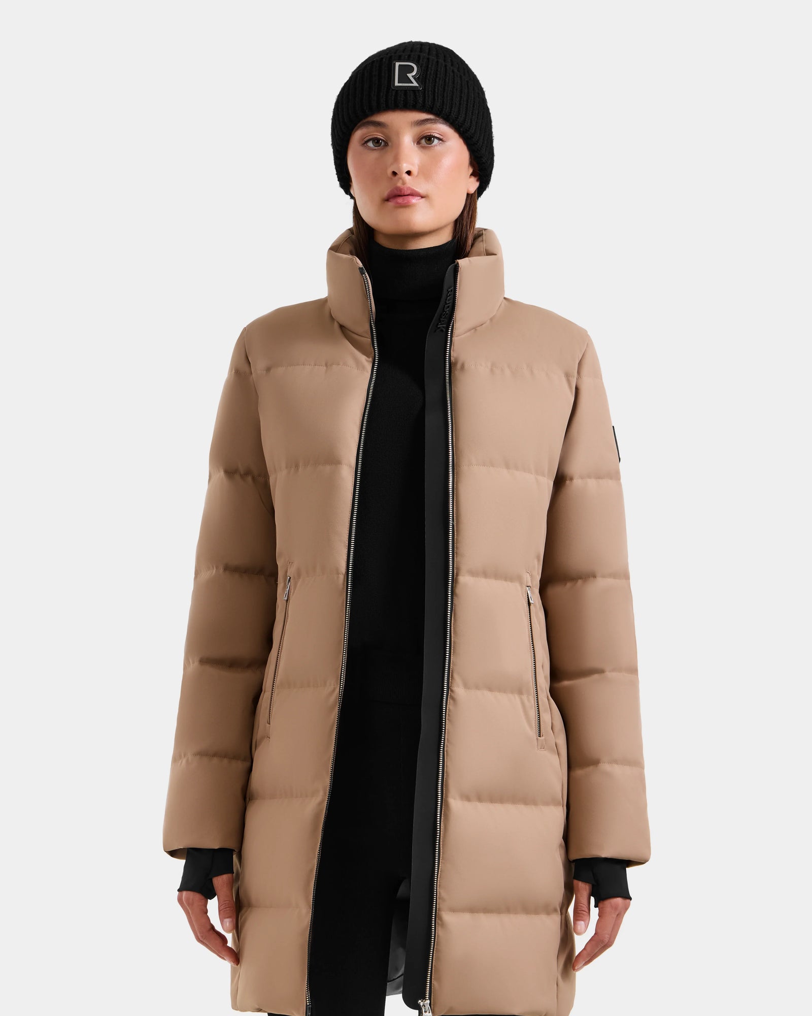 A person faces forward in the Rudsak MIRIAM - L123544 Sand women's down puffer coat with a high collar and front zipper, paired with a black beanie featuring a white R, against a plain light gray background.