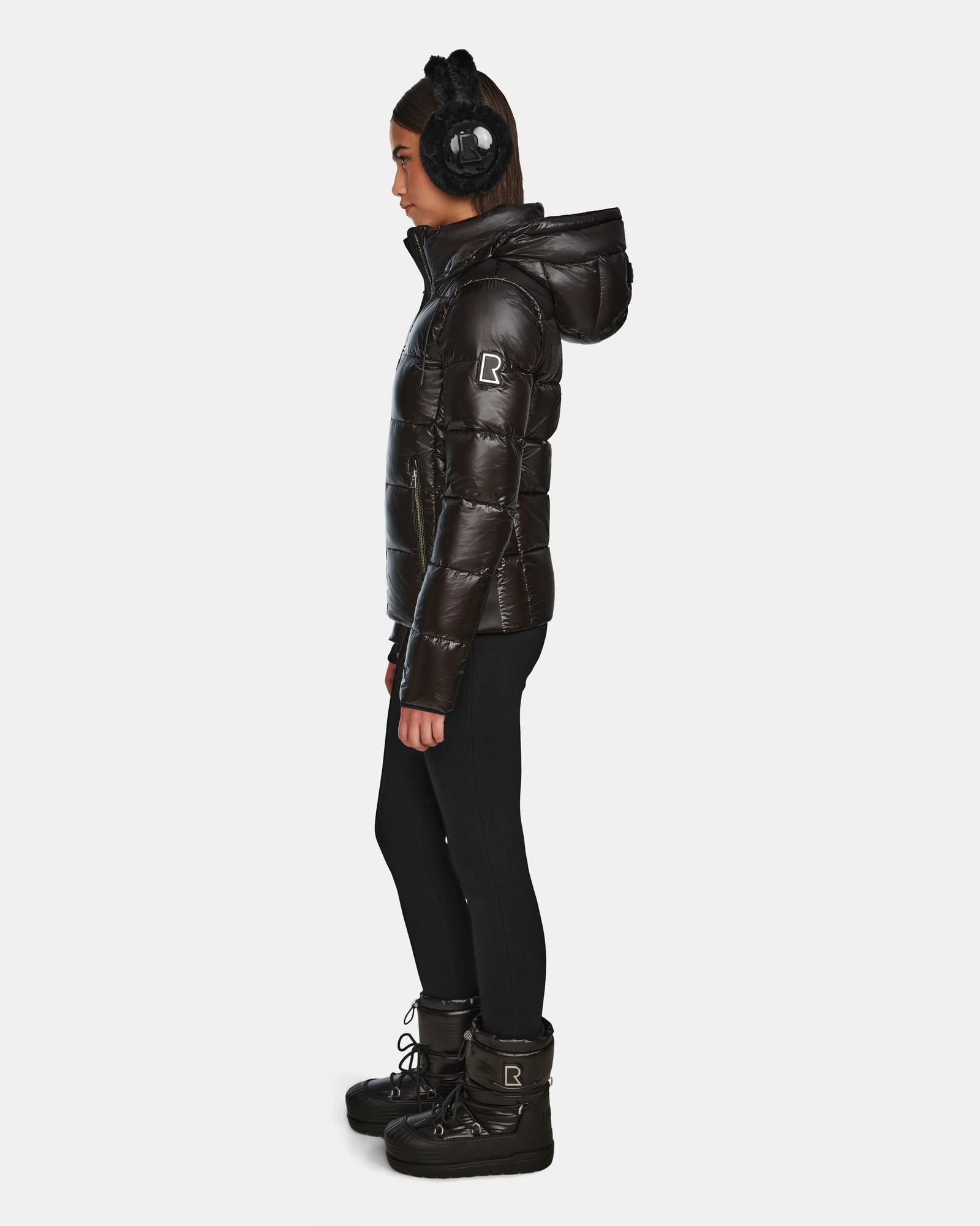 A woman in profile wears black earmuffs, black leggings, and winter boots, showcasing the Rudsak MIRRY S - L123545 Black quilted down coat with hood, set against a plain white background.