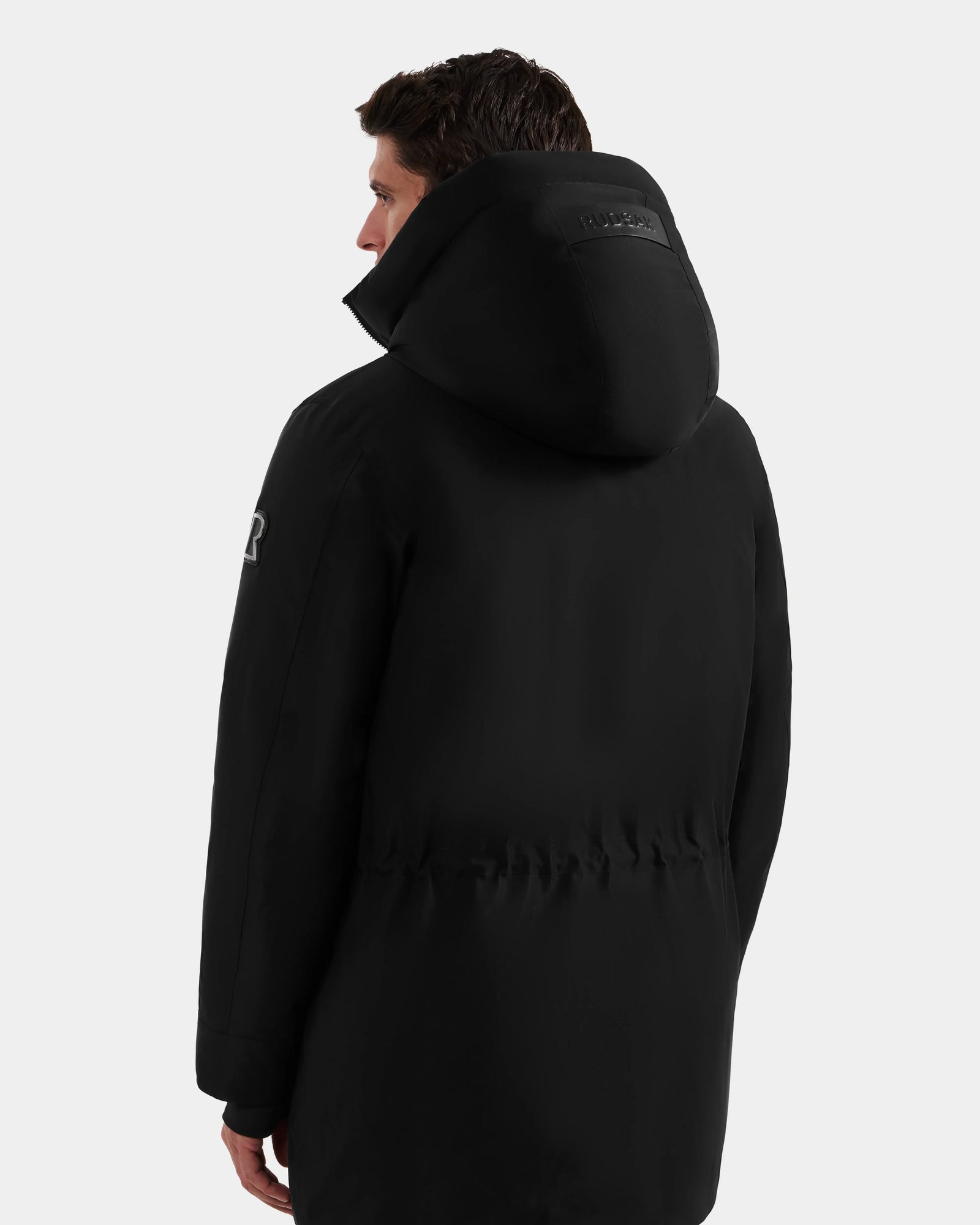 A person wearing the Rudsak MITCHELL - M125513 Black parka is shown from the back with the hood up, head turned slightly left, against a plain light background.