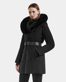 A woman with straight brown hair and a neutral expression models the Rudsak MODA M - L125555 Black long belted winter coat with a large faux fur collar against a light gray background, her left hand in the pocket—ideal for winter layering.