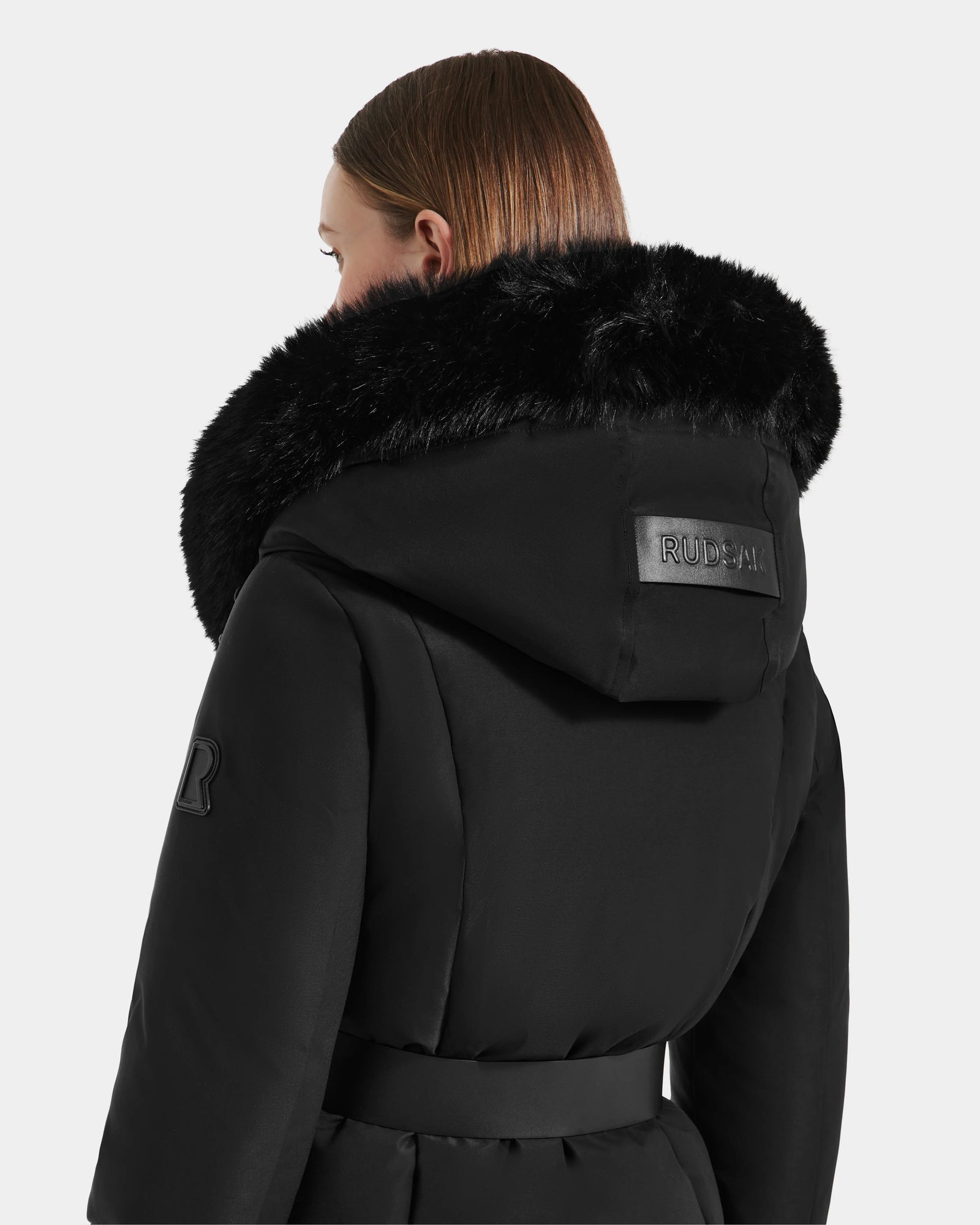 A person with straight, light brown hair wears the Rudsak MODA M - L125555 Black down puffer coat, featuring a large faux fur hood with a RUDSAK patch. The plain, light background highlights its cozy winter style.