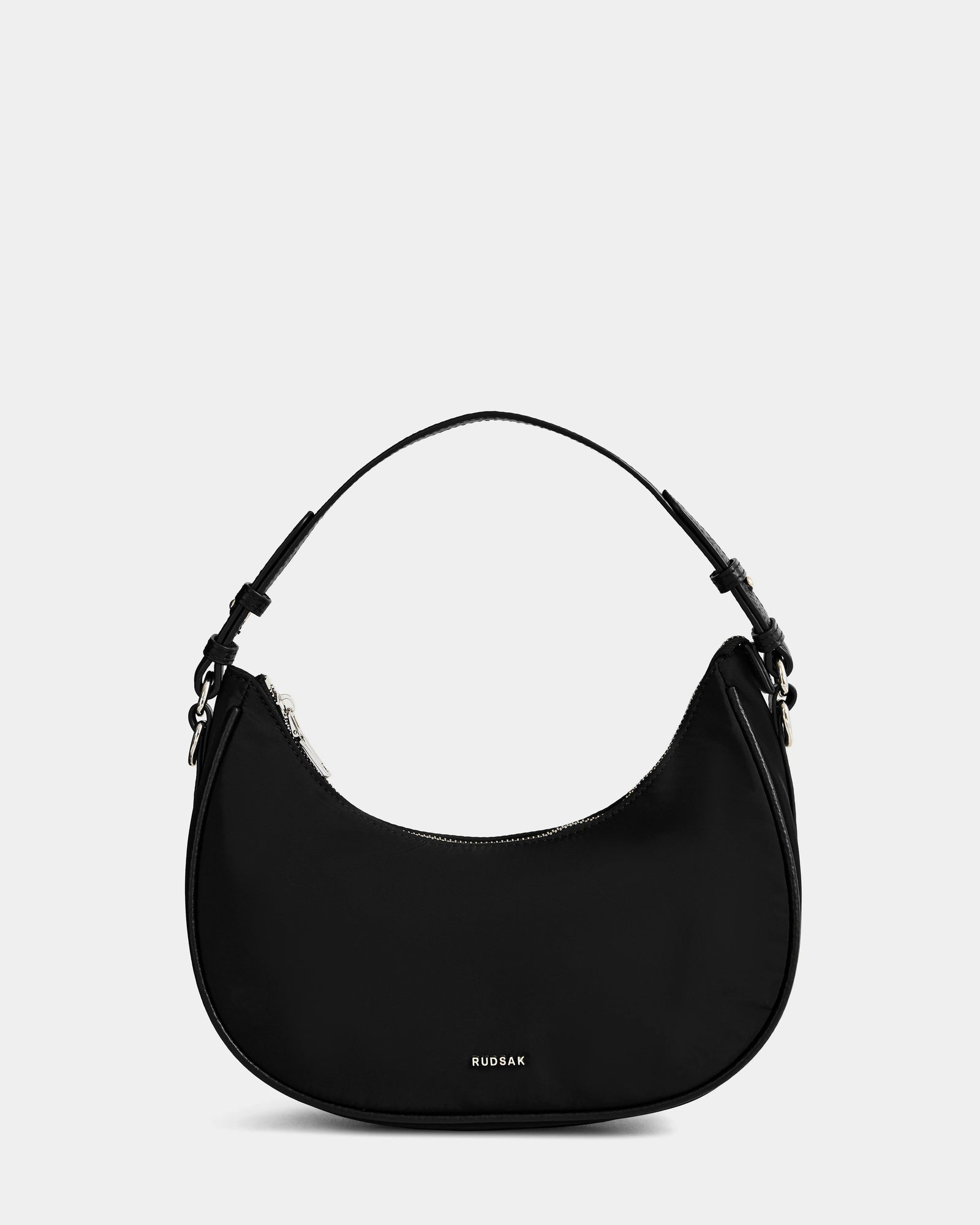 The Rudsak NIKKI - L325512 Black is a women’s crescent hobo shoulder bag with a short strap, silver zipper, and small front “RUDSAK” logo, shown against a plain light gray background.