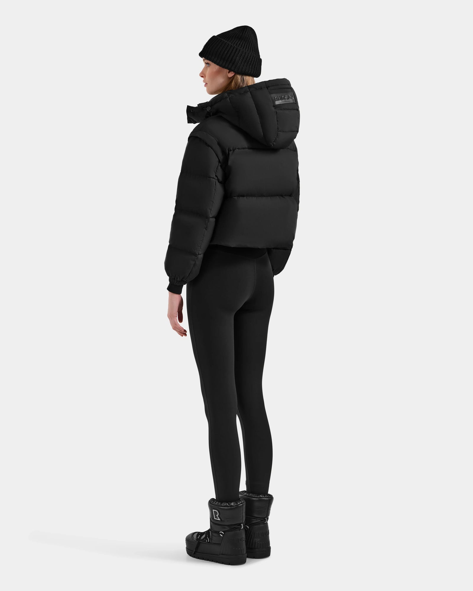 A person stands facing away, wearing the Rudsak OPRAH - L125546 Black women’s cropped down puffer with removable sleeves, paired with a black beanie, black leggings, and winter boots against a plain light background.