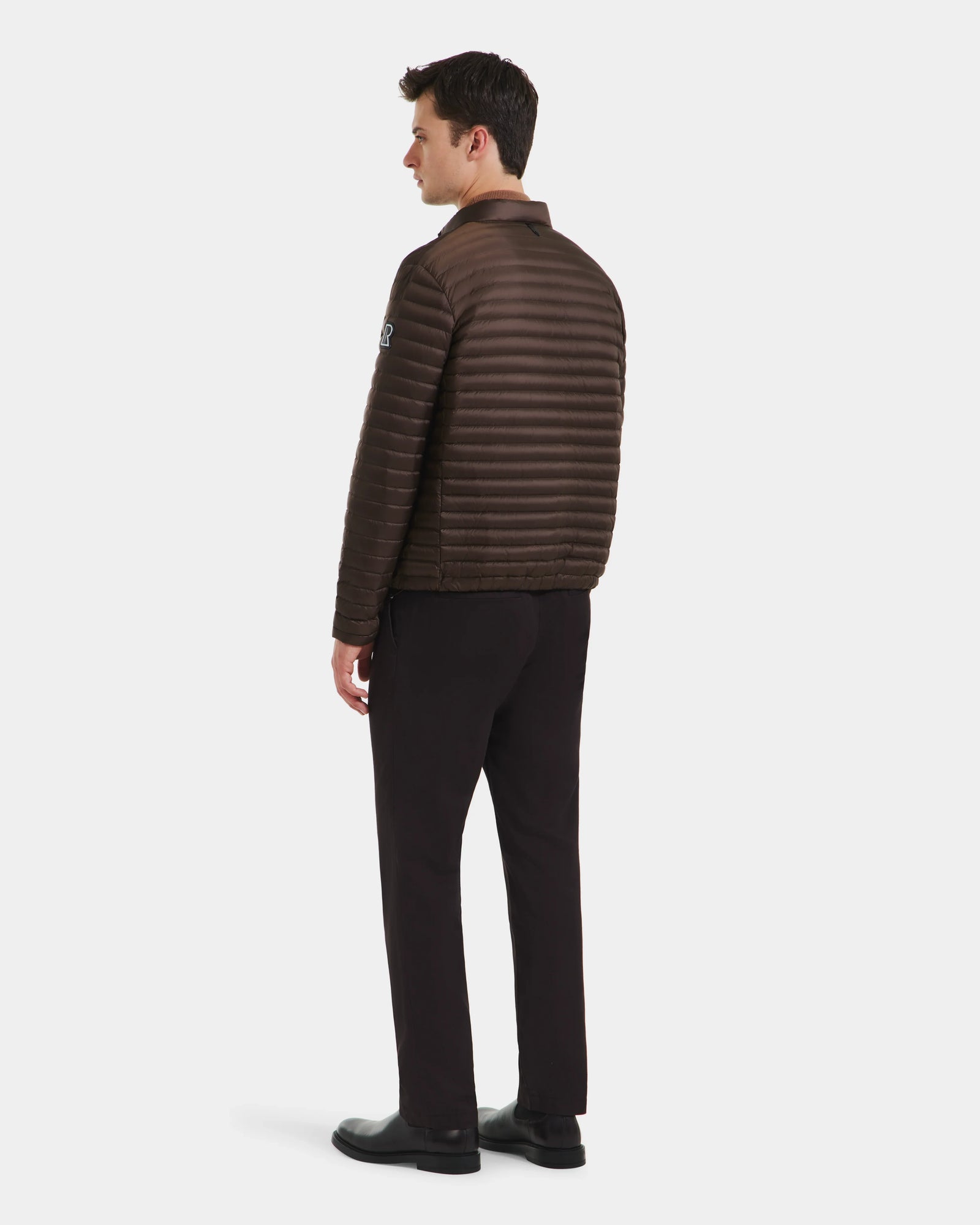 A man faces away, wearing the Rudsak REYMOND M - M125523 Mocha brown quilted puffer men's down jacket in recycled nylon, paired with black pants and shoes, against a plain white background.