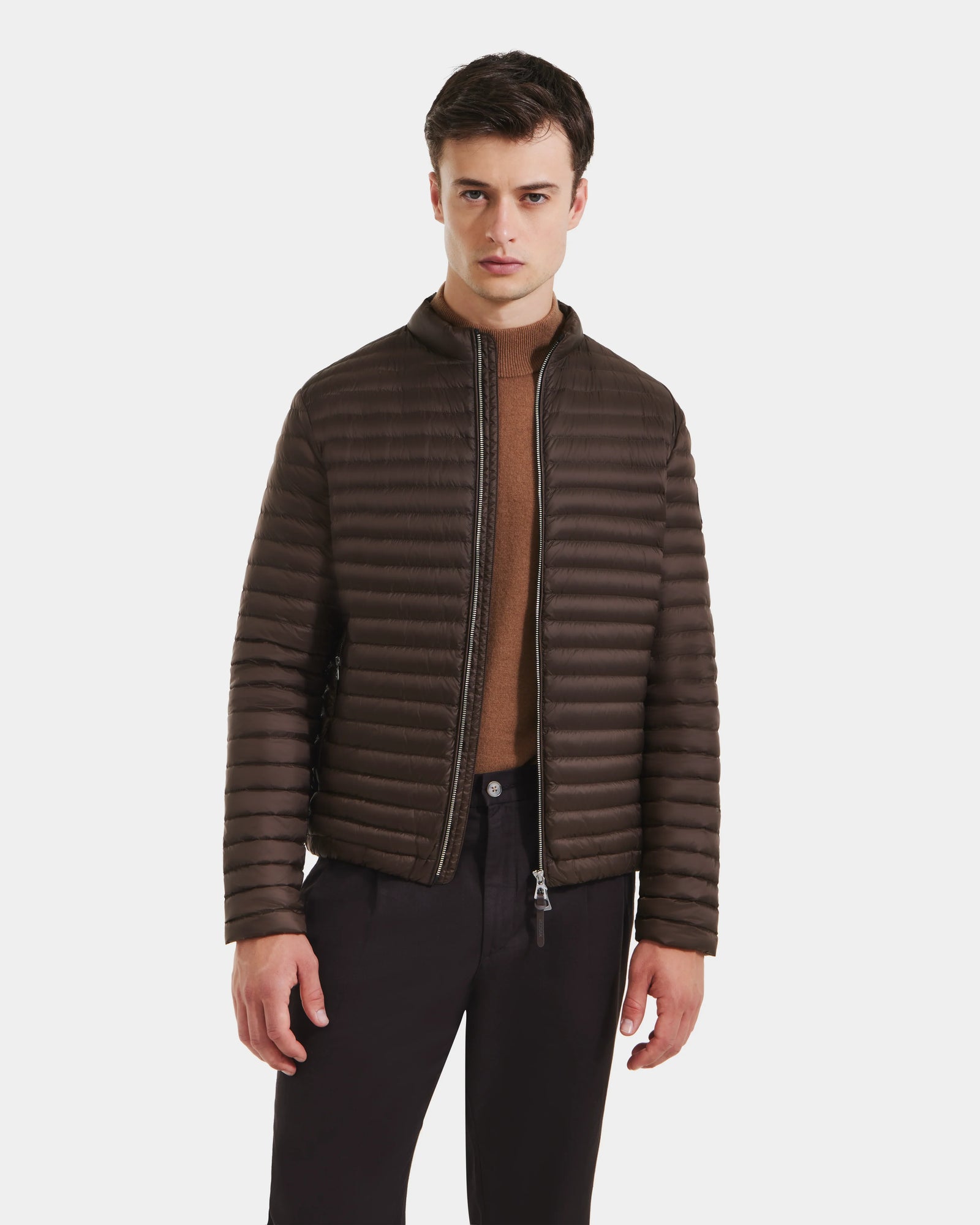 A man wearing the Rudsak REYMOND M - M125523 Mocha dark brown, RDS-certified down puffer jacket over a tan sweater and black pants stands against a plain white background.