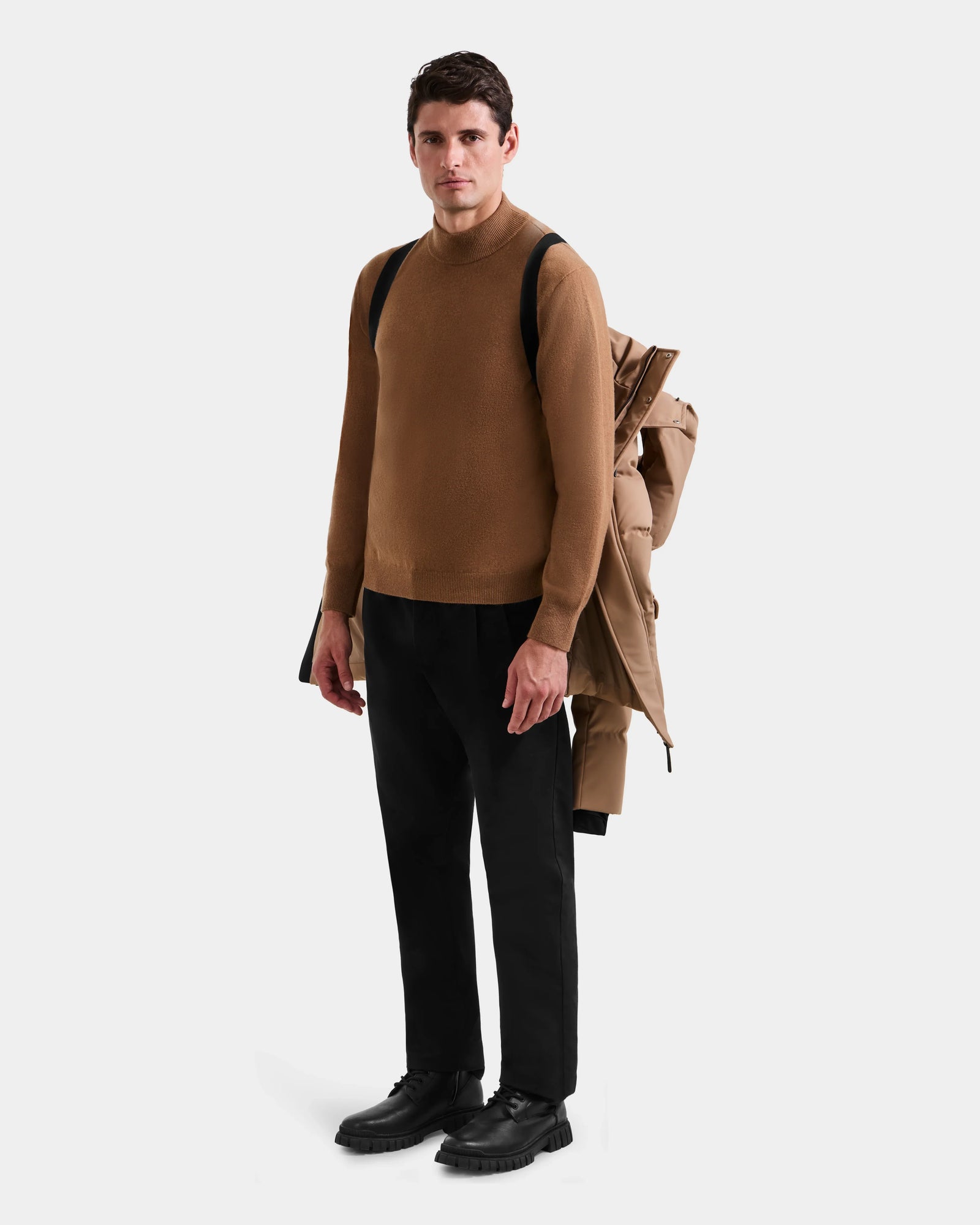 A man stands facing forward in a brown turtleneck, black pants, and black shoes. Draped over his shoulder is the Rudsak RICKY - M123502 Sand coat with a black strap—a stylish winter statement against a plain white background.