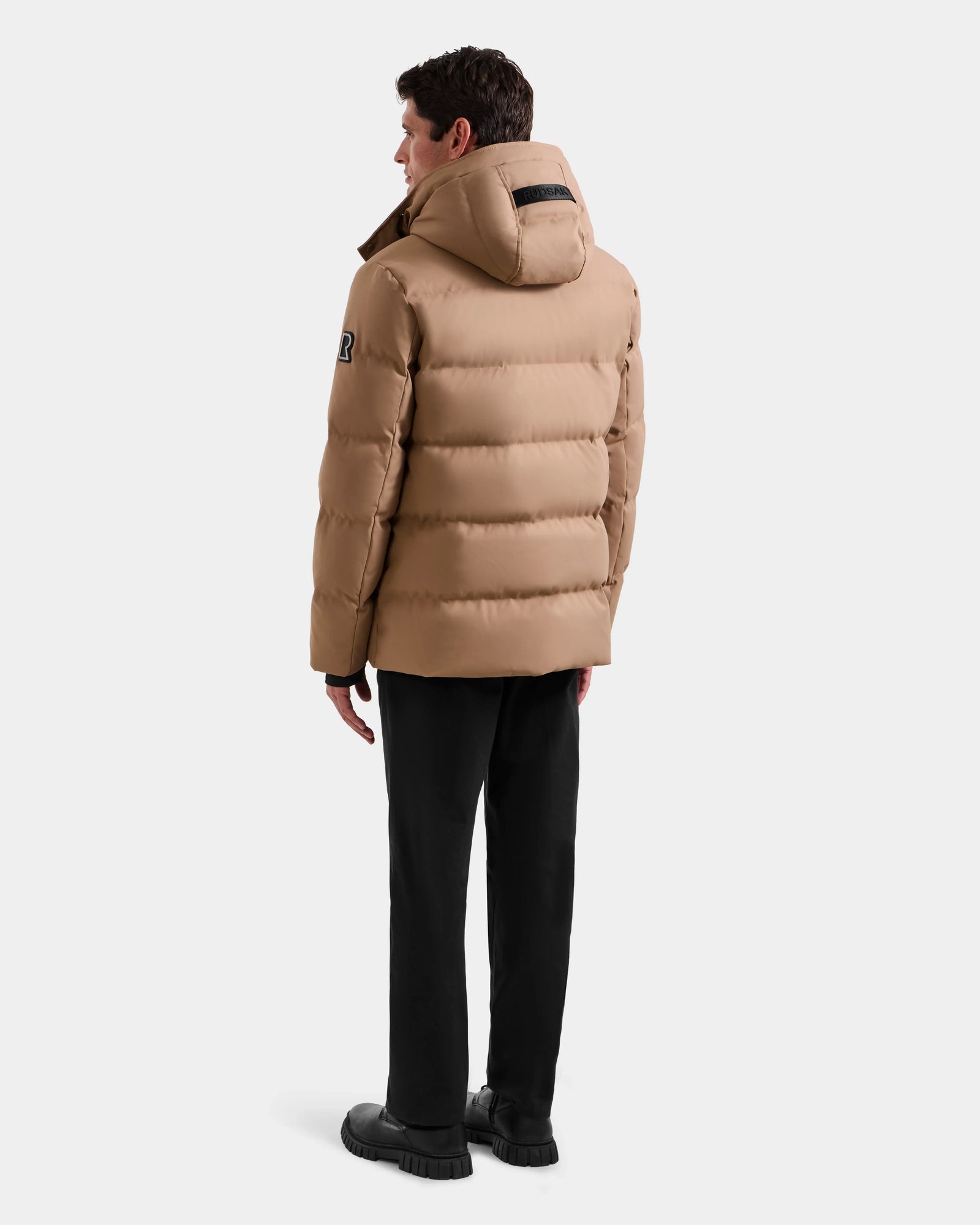 A man stands facing away, wearing the Rudsak RICKY - M123502 Sand quilted down parka with a hood, black pants, and black shoes against a plain light background—ideal for sleek winter style.