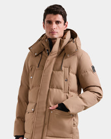 A man wears the Rudsak RICKY - M123502 Sand men's quilted down parka, featuring a hood, front pockets, and black drawstrings, against a plain light background—an ideal choice for winter fashion.