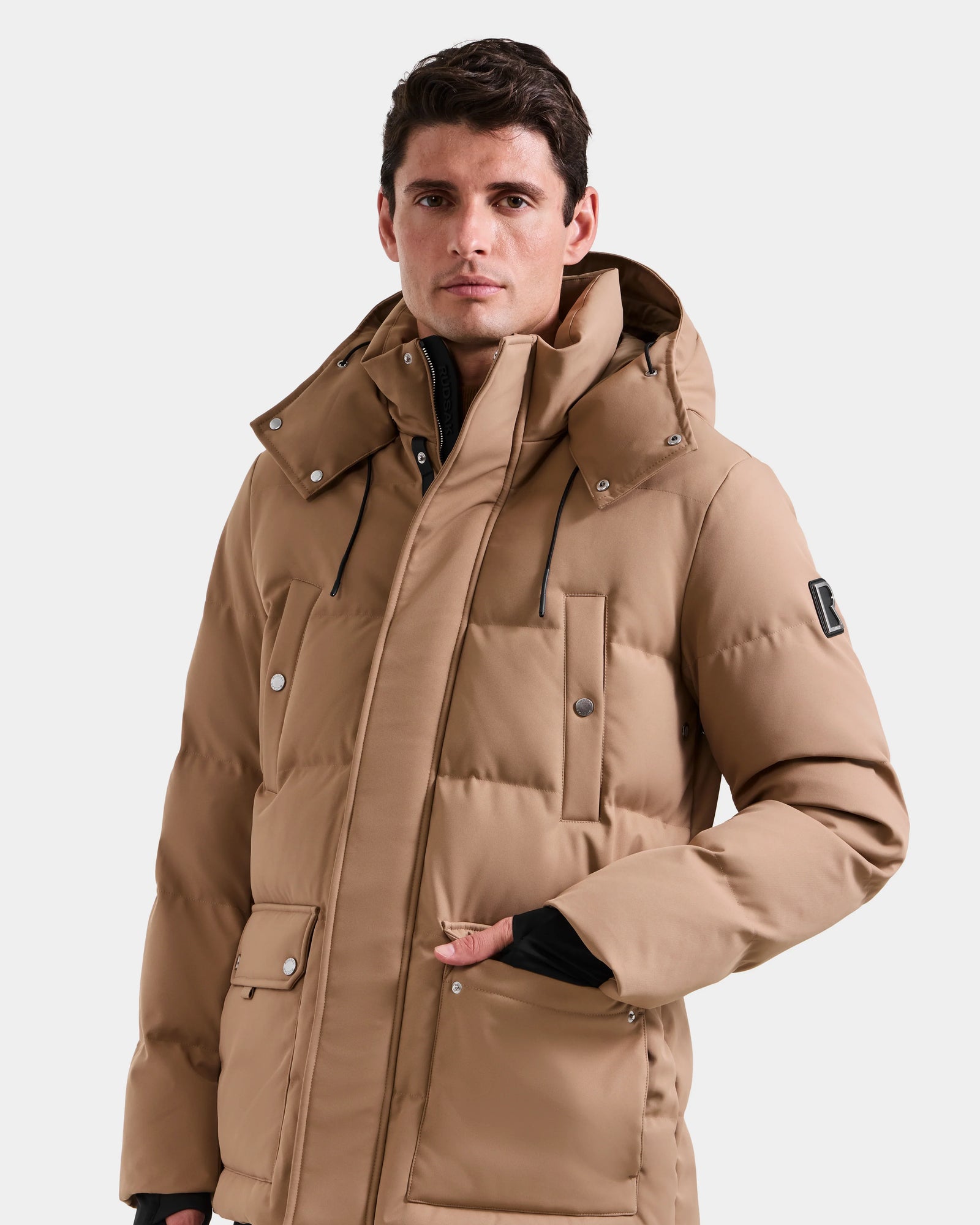 A man wears the Rudsak RICKY - M123502 Sand men's quilted down parka, featuring a hood, front pockets, and black drawstrings, against a plain light background—an ideal choice for winter fashion.