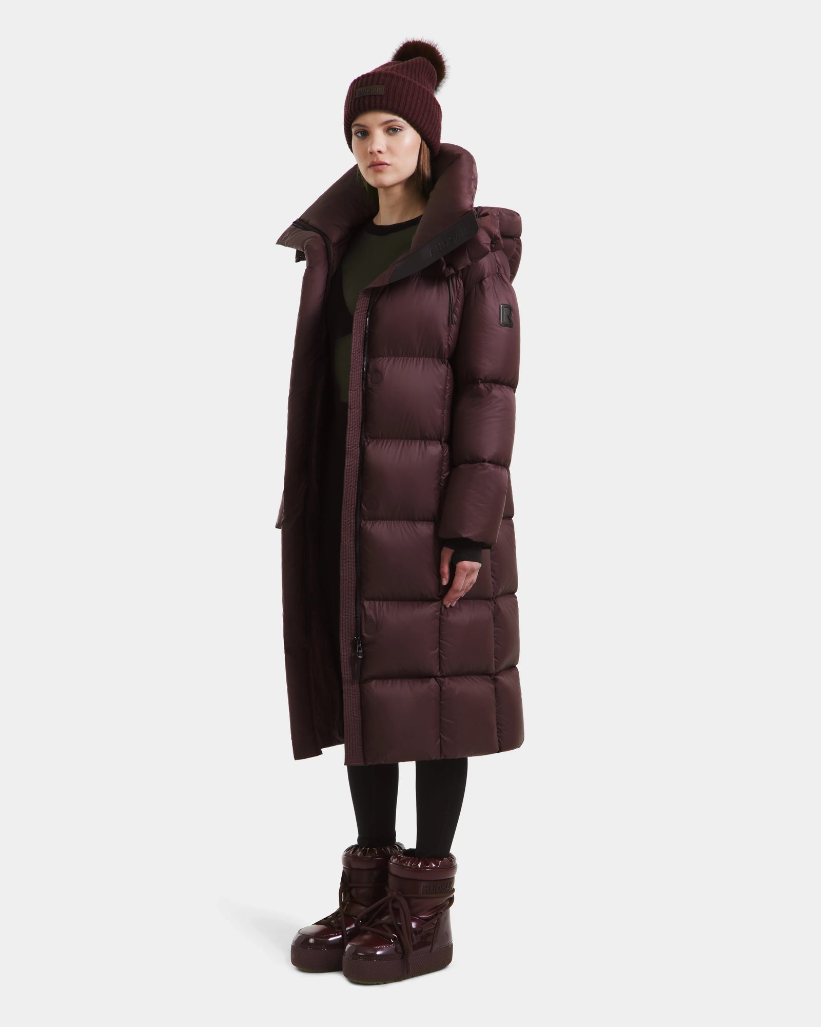 A woman wears the Rudsak SASHA - 8125686 Bordeaux women’s long down puffer coat with a high collar, paired with a matching beanie, black leggings, and shiny maroon boots, set against a plain white background.