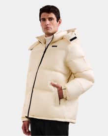 A man models the Rudsak STEFANO - 6125574 Vanilla, a cream quilted hooded jacket with a black zipper, standing against a light gray background—an ideal winter outerwear choice.