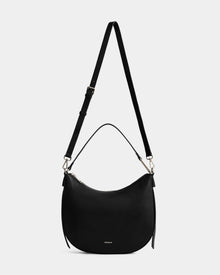 The Rudsak SYLVIA - L325515 Black is a women's leather crescent-shaped shoulder bag with an adjustable long strap, short handle, gold hardware, and a subtle embossed logo on the front, shown against a white background.