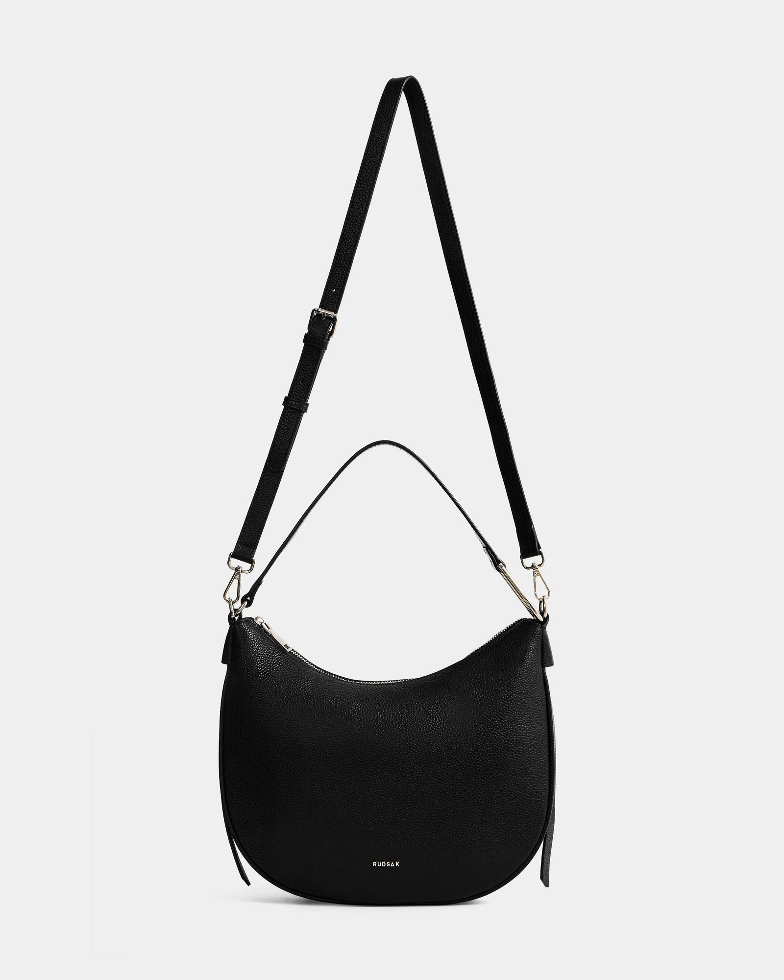 The Rudsak SYLVIA - L325515 Black is a women's leather crescent-shaped shoulder bag with an adjustable long strap, short handle, gold hardware, and a subtle embossed logo on the front, shown against a white background.