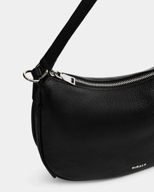 Close-up of the Rudsak SYLVIA - L325515 Black crossbody bag, crafted in pebbled leather with a silver zipper, small metallic logo, shoulder strap, and crescent silhouette.