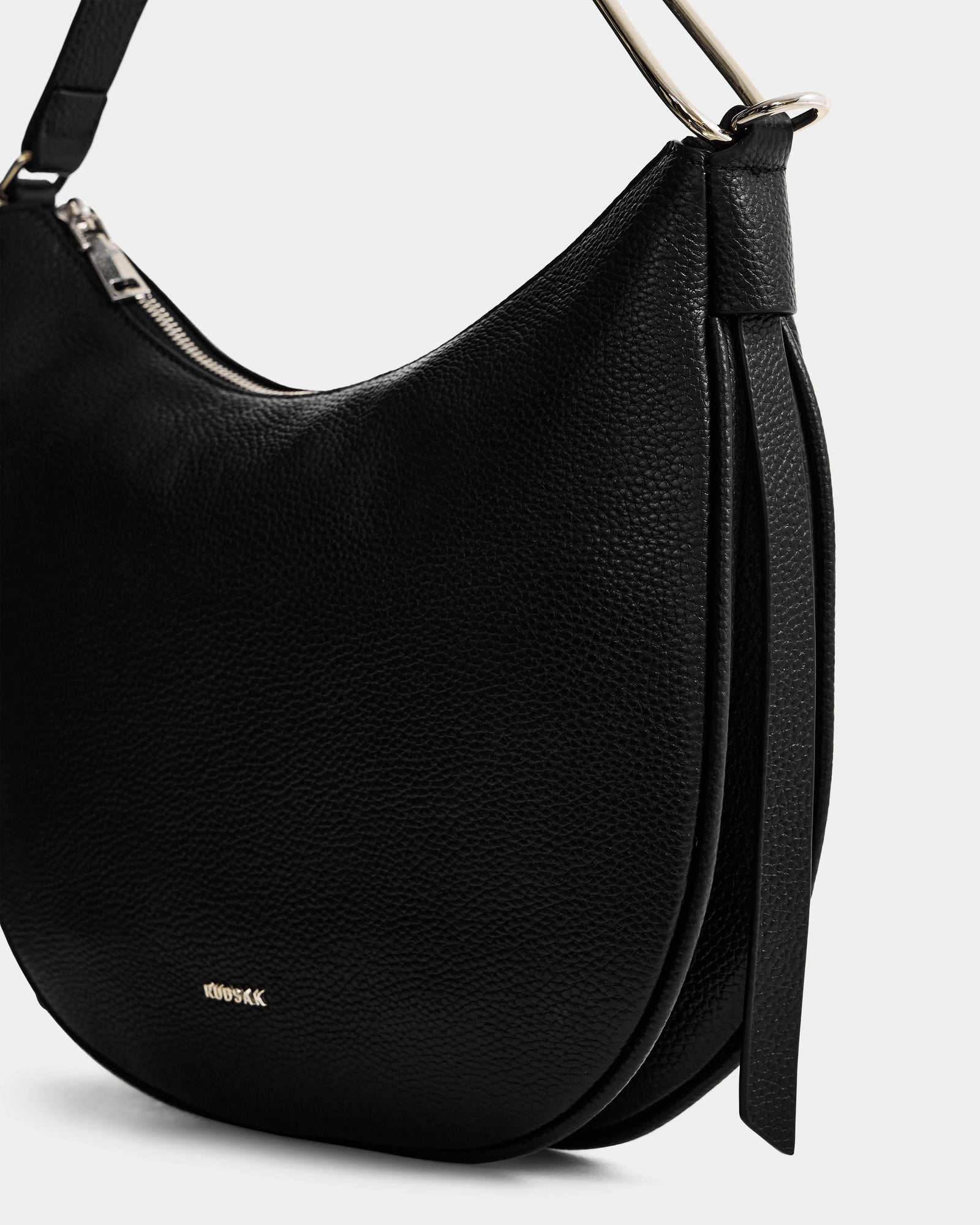 Close-up of the SYLVIA - L325515 Black crescent silhouette bag by Rudsak, featuring black textured leather, silver hardware, a zipper closure, and a strap with a decorative leather tie. The perfect chic women’s shoulder bag.