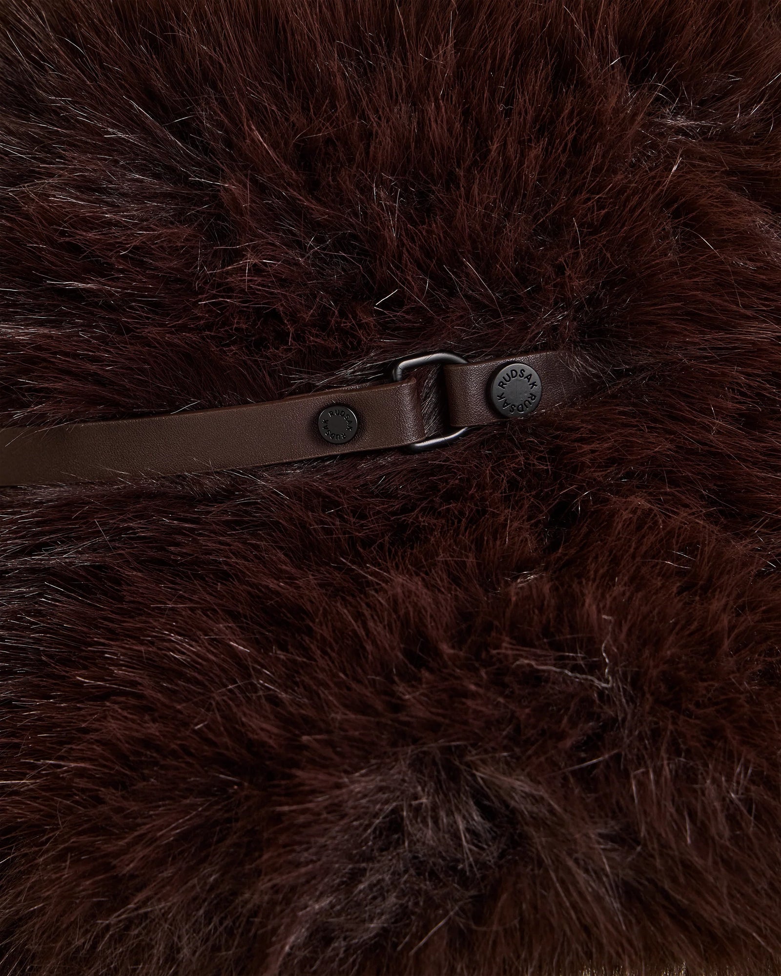 A close-up of the YURI - L425538 Espresso by Rudsak: dark brown fluffy faux fur, brown leather strap, metallic buckle and button fastener—this stylish bucket hat is a perfect winter accessory.