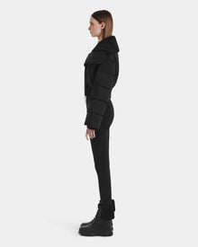 A woman stands in profile against a plain background, wearing the Rudsak YVETTE - L125557 Black women’s cropped down jacket with an oversized collar, paired with black leggings and black lace-up boots with fur trim.