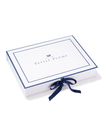 A rectangular white box features Petite Plume centered, bordered by a thin blue line, adorned with a blue bow and a small bird icon above the text. Perfect for encasing Kids Delphine Slipper in Black Velvet, it offers luxurious comfort for pampering little feet.