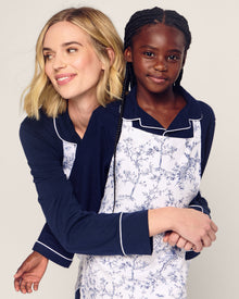 A blonde woman in a navy and white outfit embraces a young girl with braids. Both wear Petite Plumes Kids Apron in Timeless Toile, ready for holiday baking, their smiles brightening the plain backdrop with festive joy.