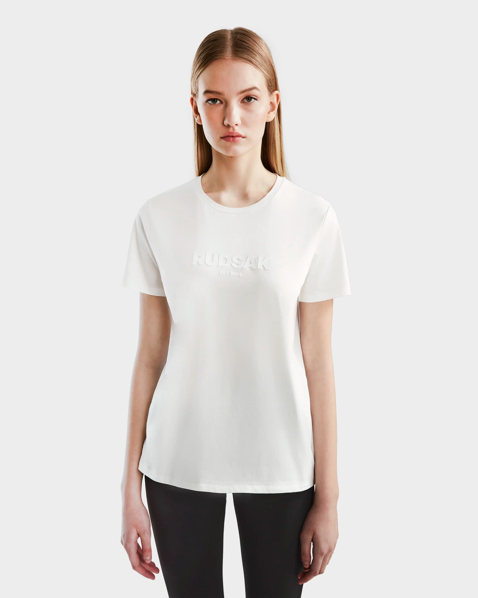 A young woman with straight, long blonde hair wears the Rudsak AMINA - 8524012 Cotton White women’s crewneck short sleeve t-shirt with a subtle front embossing, paired with black pants, standing against a plain white background.
