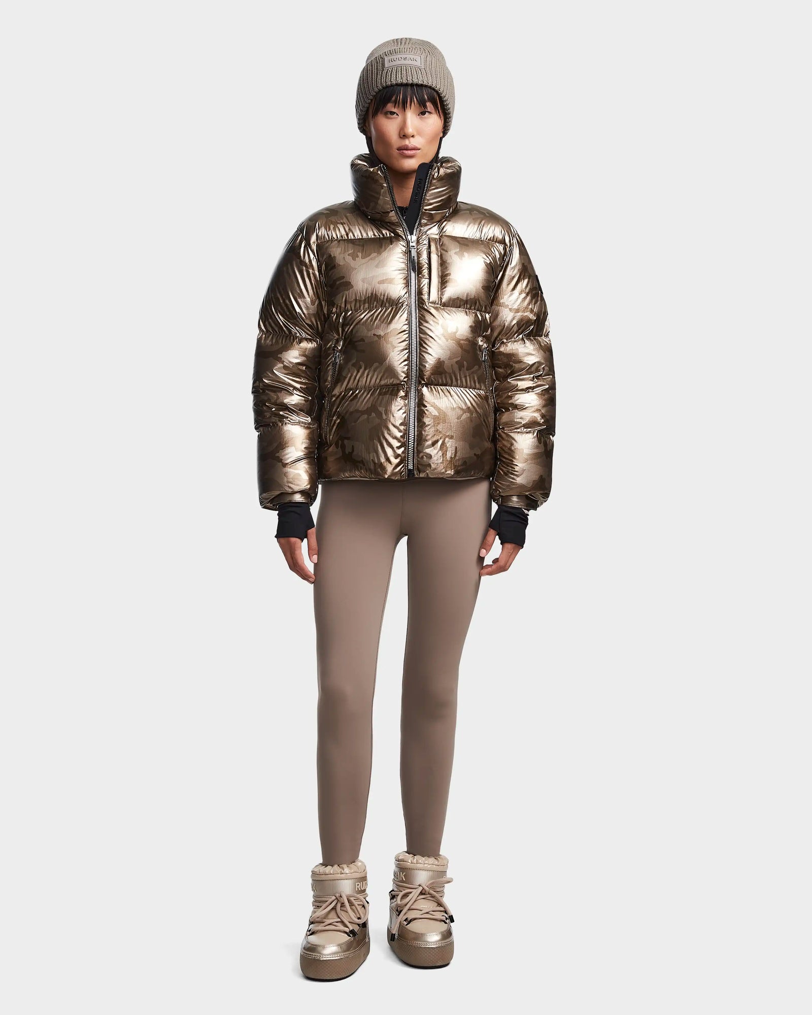 A person wears the Rudsak MAVE S - 8123563 Metallic Ivory Camo women’s down puffer jacket, taupe leggings, tan winter boots, and a gray knit beanie, standing facing forward against a plain light background.
