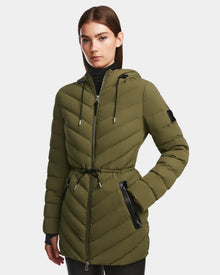 A woman wears the Rudsak FRANCINE - 8124722 Loden Green puffer jacket with black drawstrings and zippered pockets, standing with one hand in her pocket against a plain light background.