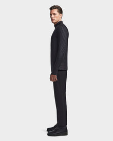A man stands in profile against a plain background, wearing the Rudsak WARREN - 6523502 Black Pattern mens fleece-lined fitted stretch top, paired with black pants and shoes.