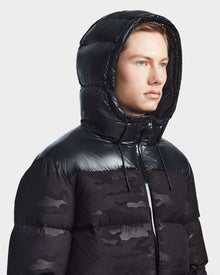 Wearing the Rudsak RONNY M - M123512 Camo Black, a shiny black hooded down puffer with a subtle camo pattern, this stylish and weather-resistant mens outerwear stands out against a plain light background.
