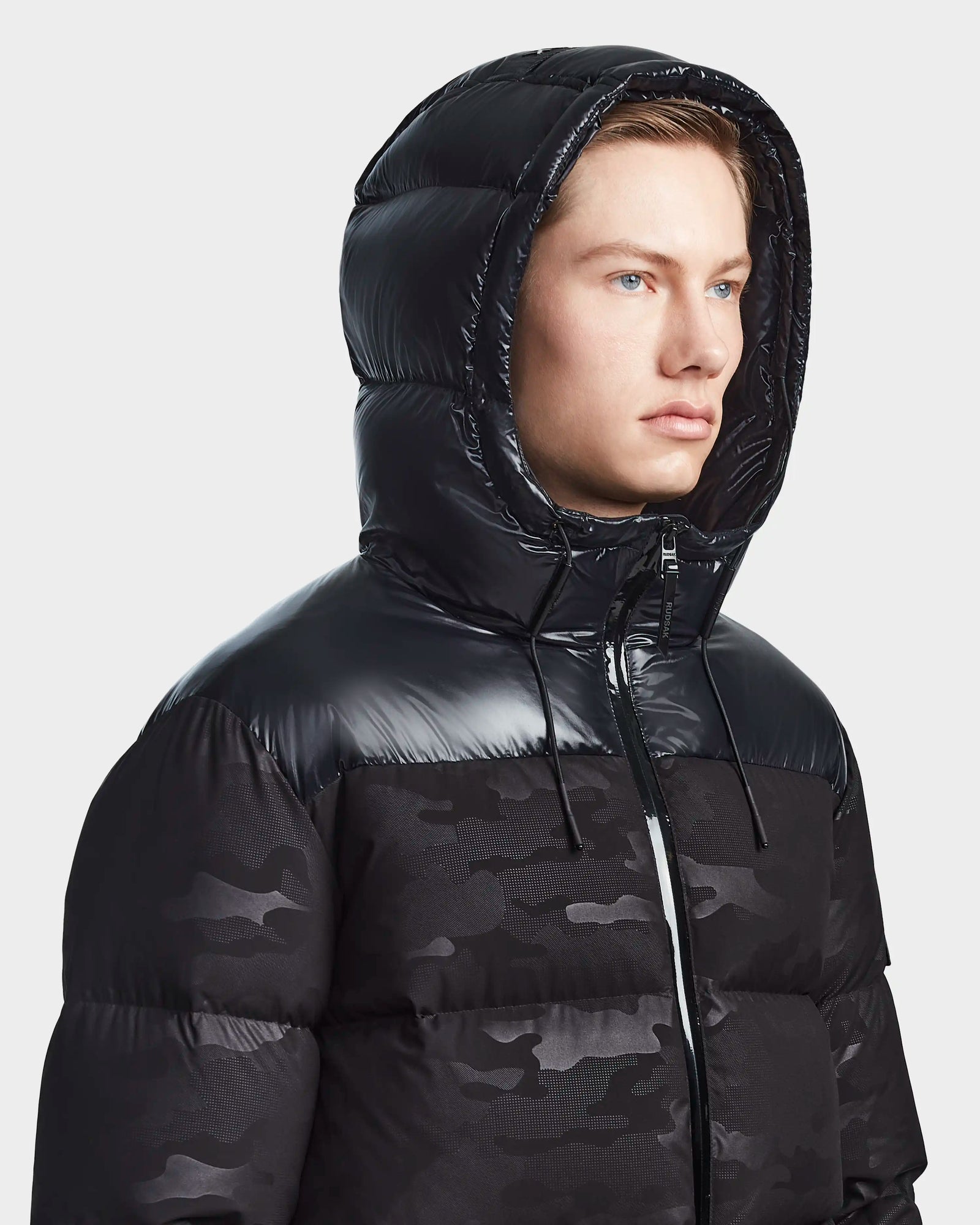 Wearing the Rudsak RONNY M - M123512 Camo Black, a shiny black hooded down puffer with a subtle camo pattern, this stylish and weather-resistant mens outerwear stands out against a plain light background.